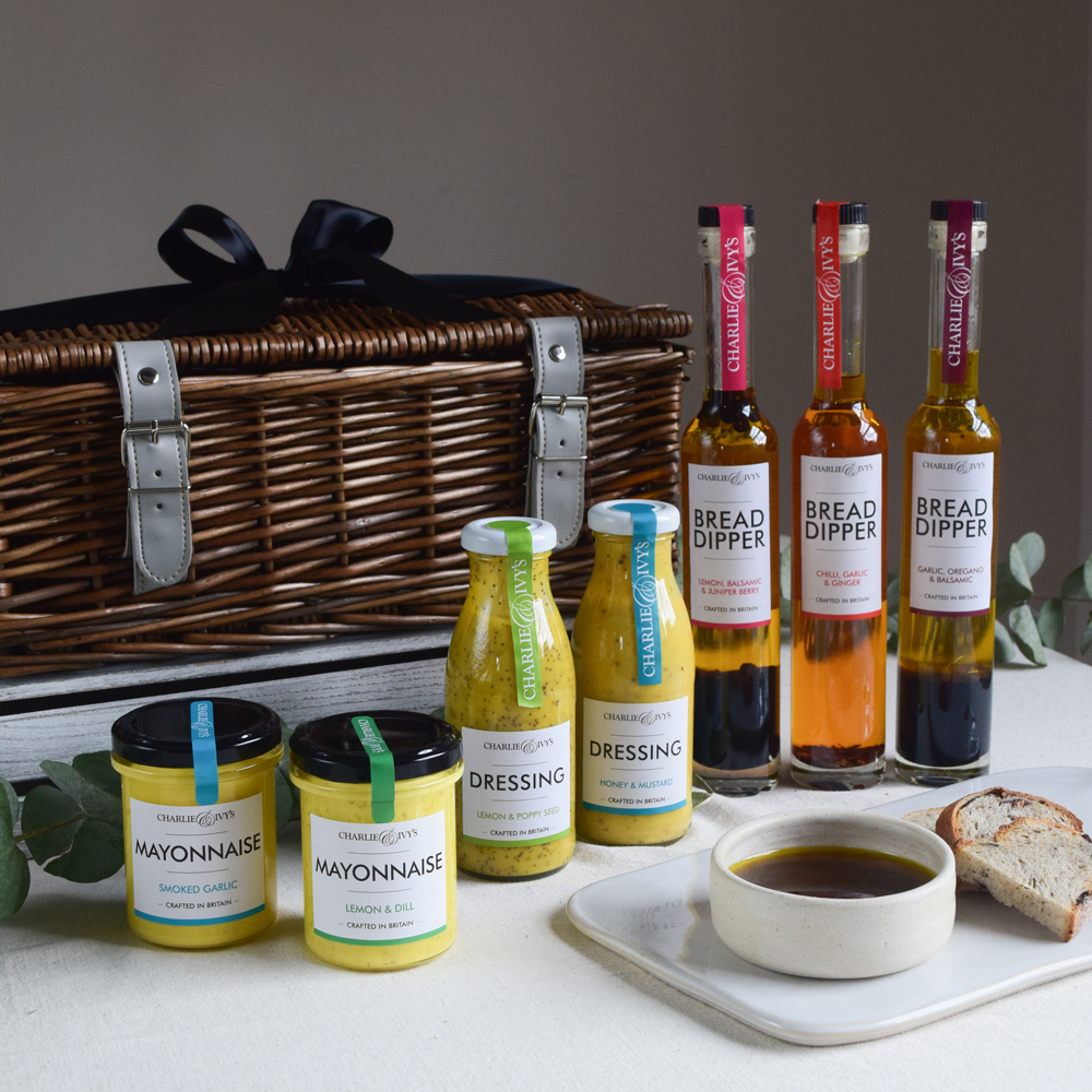A grey wicker hamper on a table top with the Charlie & Ivy's products, bread and a dipping bowl filled with oil.